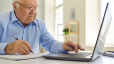 older man writing, using laptop