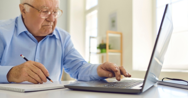 older man writing, using laptop